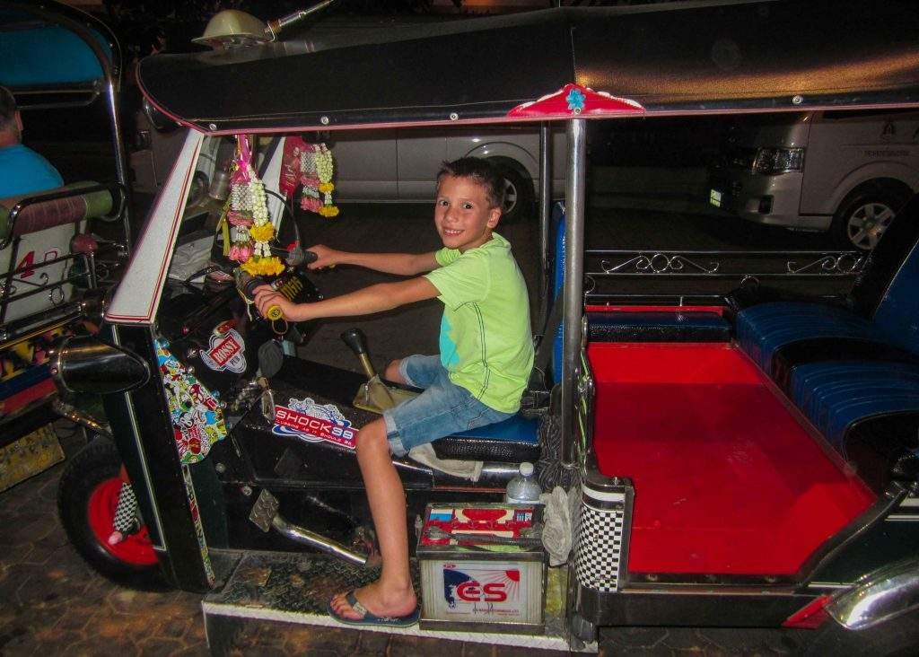 An 8 year old boy has a huge smile on his face, sitting behind the wheel of a parked red Tuk Tuk in Bangkok Thailand