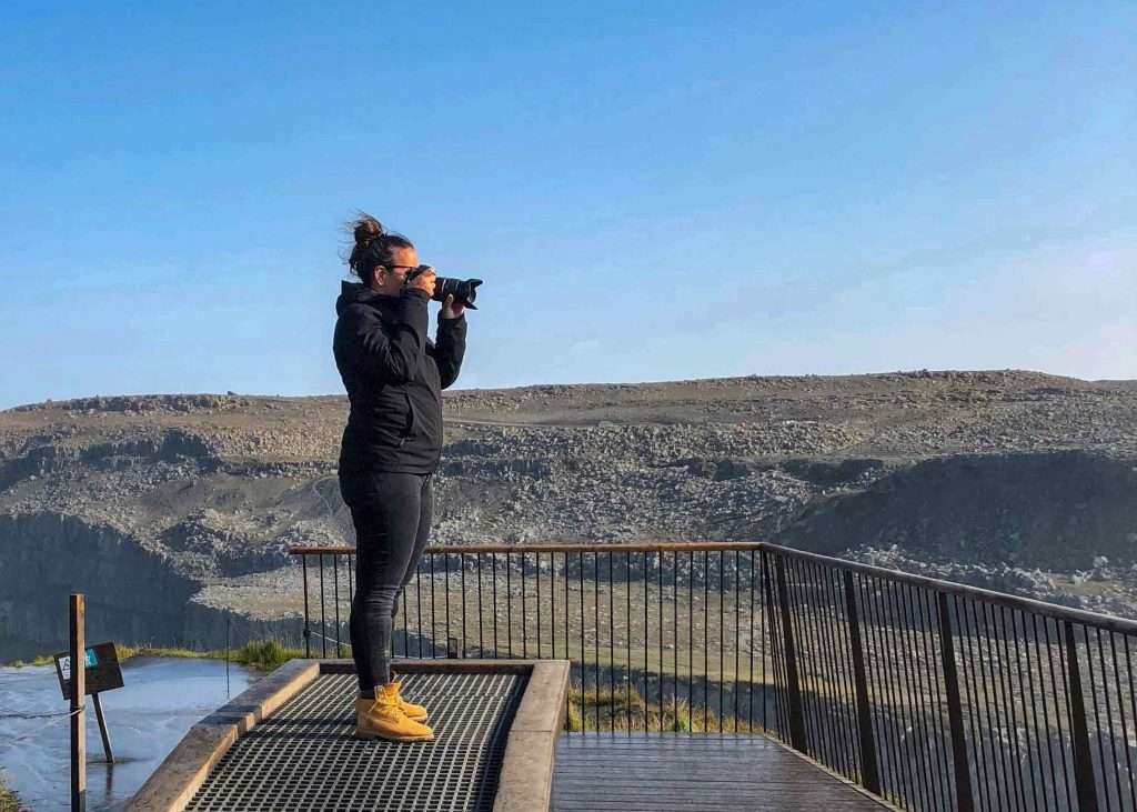 Woman in a black raincoat, black jeans and brown Timberland boots takes a picture of the Dettifoss waterfall in Iceland from a viewpoint