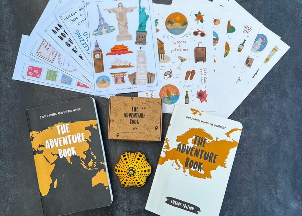 Products from The Adventure Book on a grey table. The original edition, Europe edition, wooden stamp set, printable stationery set and travel sticker set