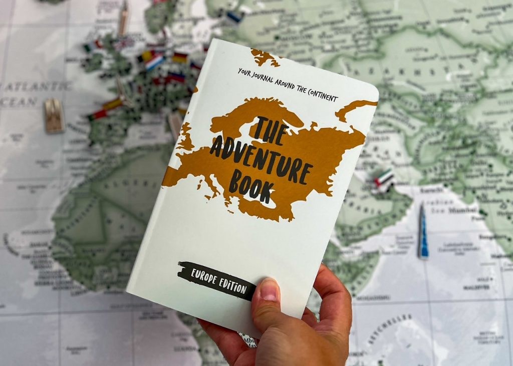 A world map filled with pins is a backdrop to a hand holding The Adventure Book - Europe edition