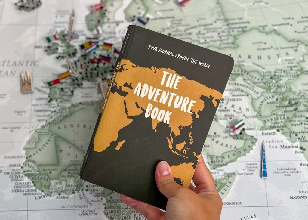 A world map filled with pins is a backdrop to a hand holding The Adventure Book - original edition