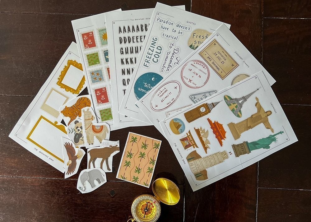 A small golden compass lies on a wooden table accompanied by stickers for a scrapbook