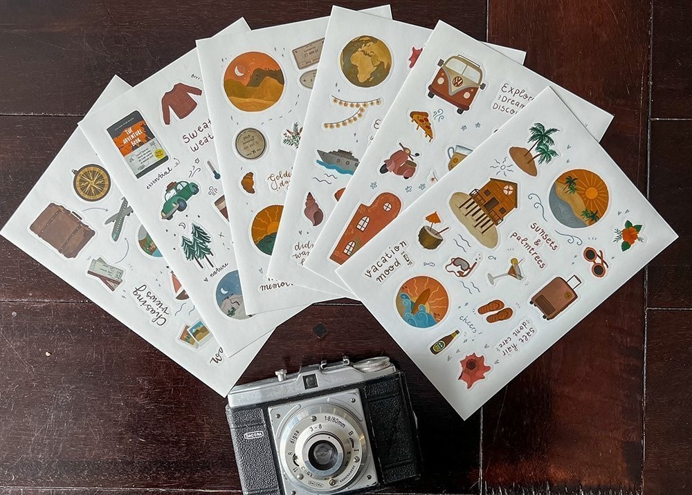 A camera lies on a wooden table accompanied by 6 sticker pages with travel stickers on it