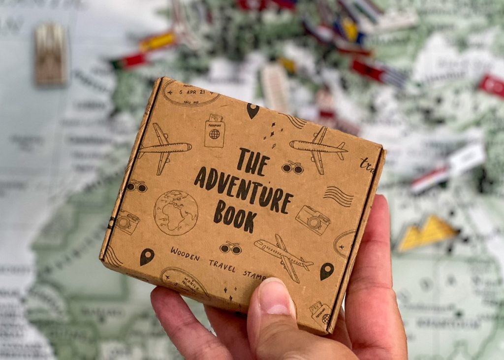 A world map filled with pins is a backdrop to a hand holding the wooden stamp set by The Adventure Book