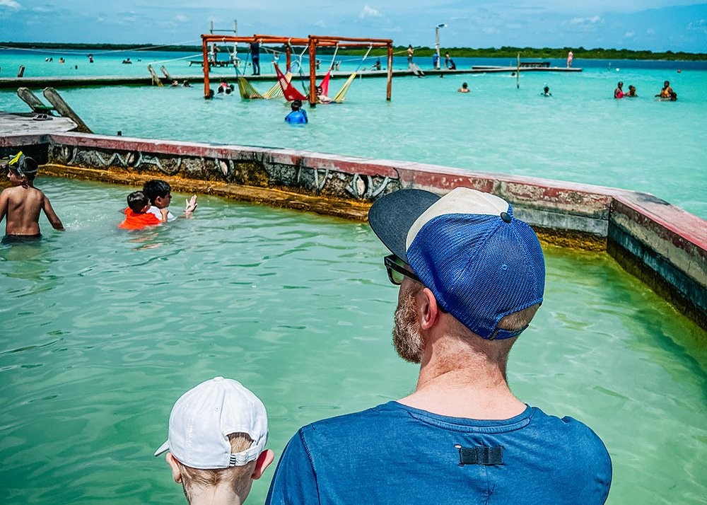 A father and son are sitting close to one another on a dock overlooking lake Bacalar. Both are wearing a cap against the sun and are admiring the turquoise color tones of this Mexican lake.